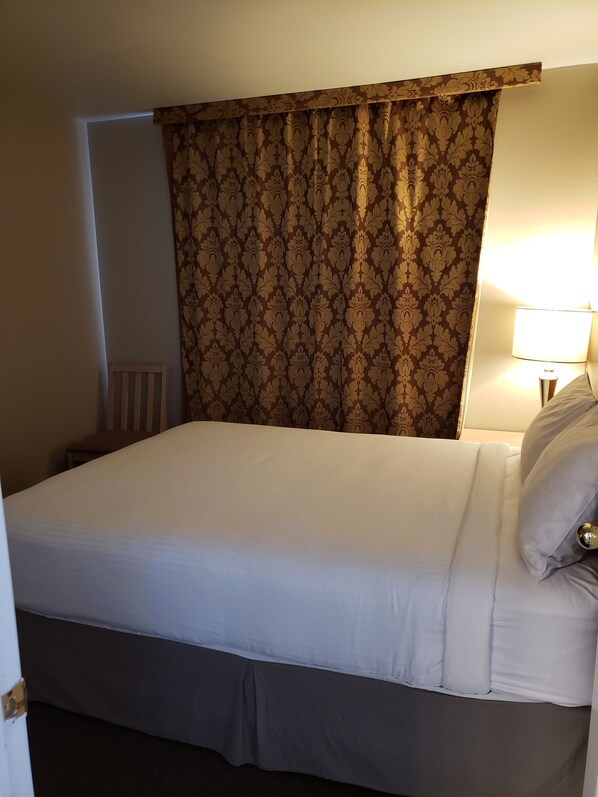 A neatly made bed with white linens, a brown patterned curtain, and a bedside lamp.