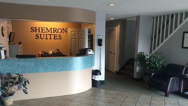 A reception area with a curved counter labeled "SHEMIRON SUITES," a staircase, and a seating area with a potted plant. - Shemron Suites Hotel (Deep River)