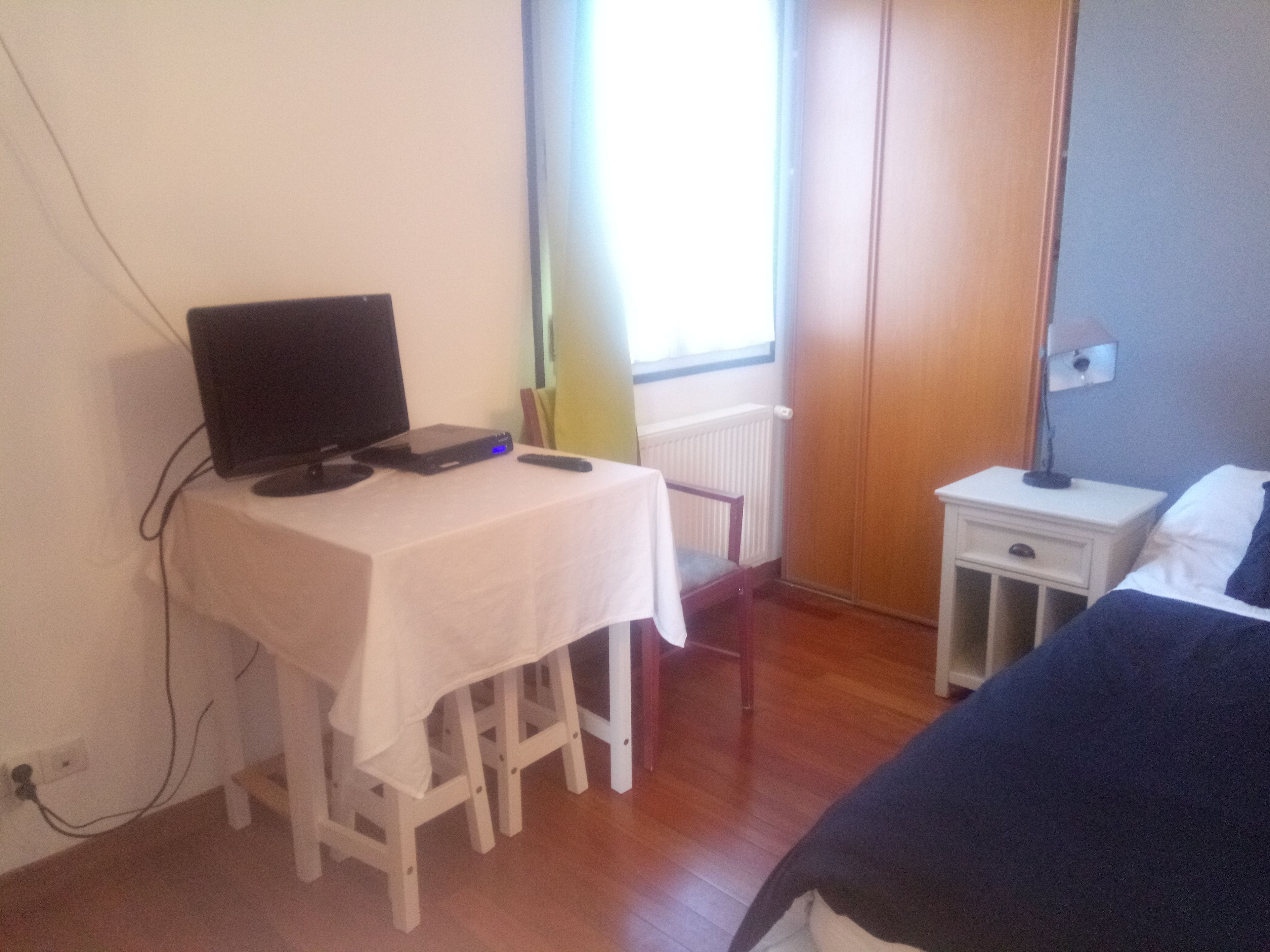 A bedroom with a bed, a desk with a computer, a chair, a nightstand, and a window with curtains.