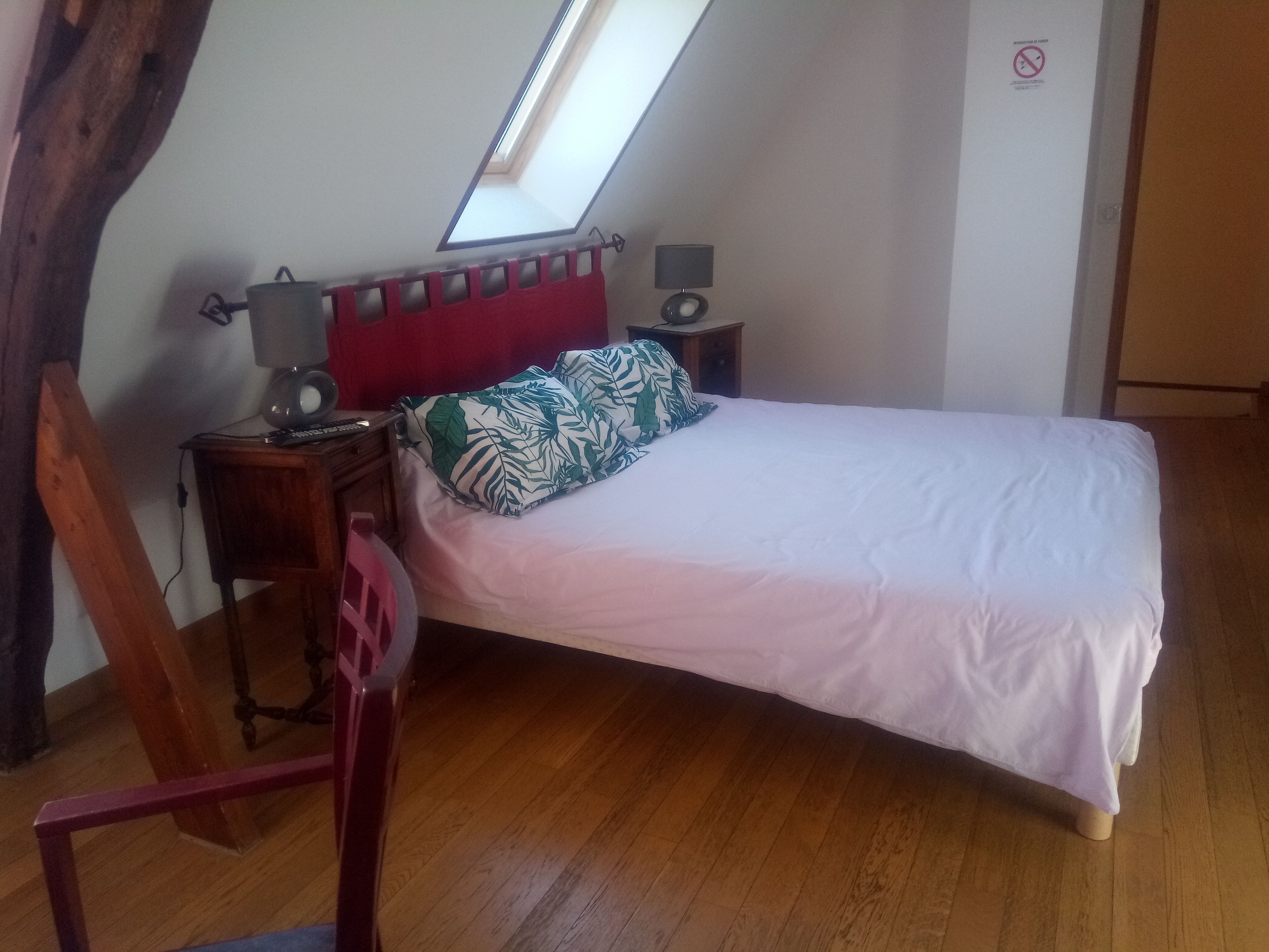 A bedroom with a sloped ceiling, a skylight, a bed with white bedding, a red headboard, a nightstand with a lamp, and a wooden chair.