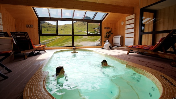 A wooden cabin with a large indoor swimming pool, wooden deck, and a scenic view of a green landscape through large windows. - Hôtel les skieurs (Bellevaux)