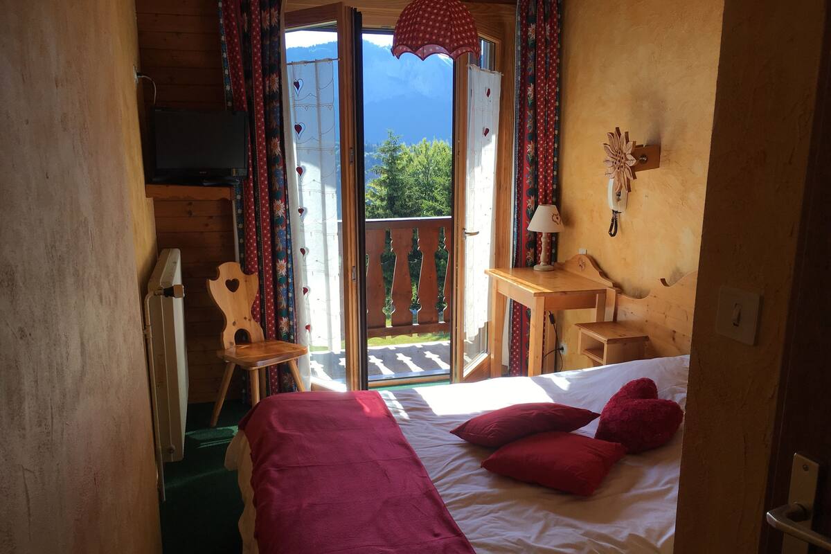 Une chambre à coucher avec un lit, des oreillers rouges, un bureau en bois, une chaise et une vue sur la verdure à l’extérieur.