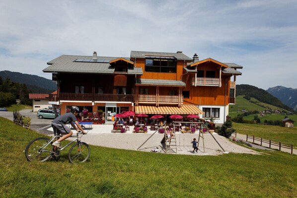 A multi-story building with a balcony, outdoor seating area, and a paved entrance. - Hôtel les skieurs (Bellevaux)