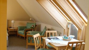 Ein kleiner, gemütlicher Dachbodenraum mit schräger Decke, einem grünen Sofa, einem Holzessttisch und einem Fenster, das für natürliches Licht sorgt.