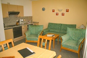 A compact kitchen with a stove, oven, and coffee maker. There is a green sofa with patterned cushions and two wooden chairs. A yellow table with a tablet is in the center. - Villa Viktoria (Loddin)