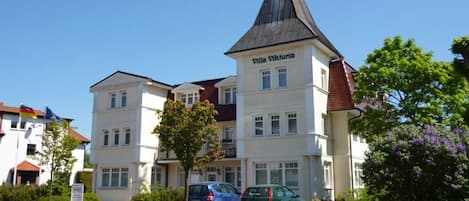 A multi-story building named Villa Viktoria with a prominent tower, surrounded by greenery and parked cars.