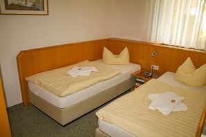 A hotel room with two beds, each with a beige bedspread and pillows. There is a wooden headboard, a bedside table with a lamp, and a small shelf with books. - Villa Viktoria (Loddin)