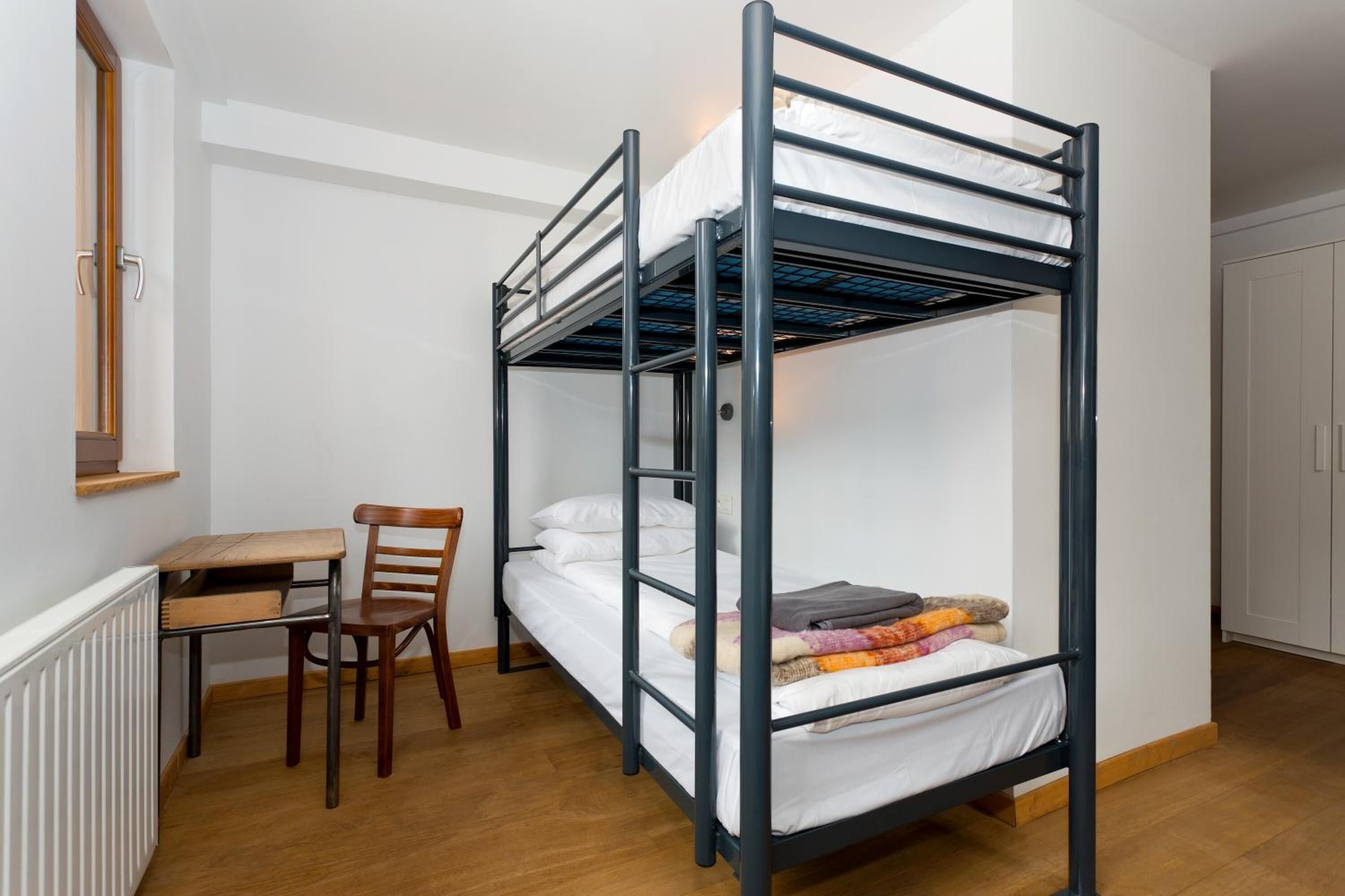Habitación con literas, escritorio, silla, paredes blancas y un radiador.