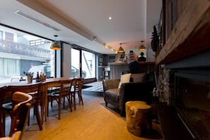 A cozy living room with a fireplace, wooden furniture, and a view of the street through large windows. - Hideout Hostel (Morzine)