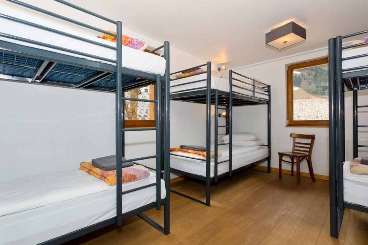 A dormitory room with bunk beds, a wooden floor, a window with a view, and a small table with a chair.