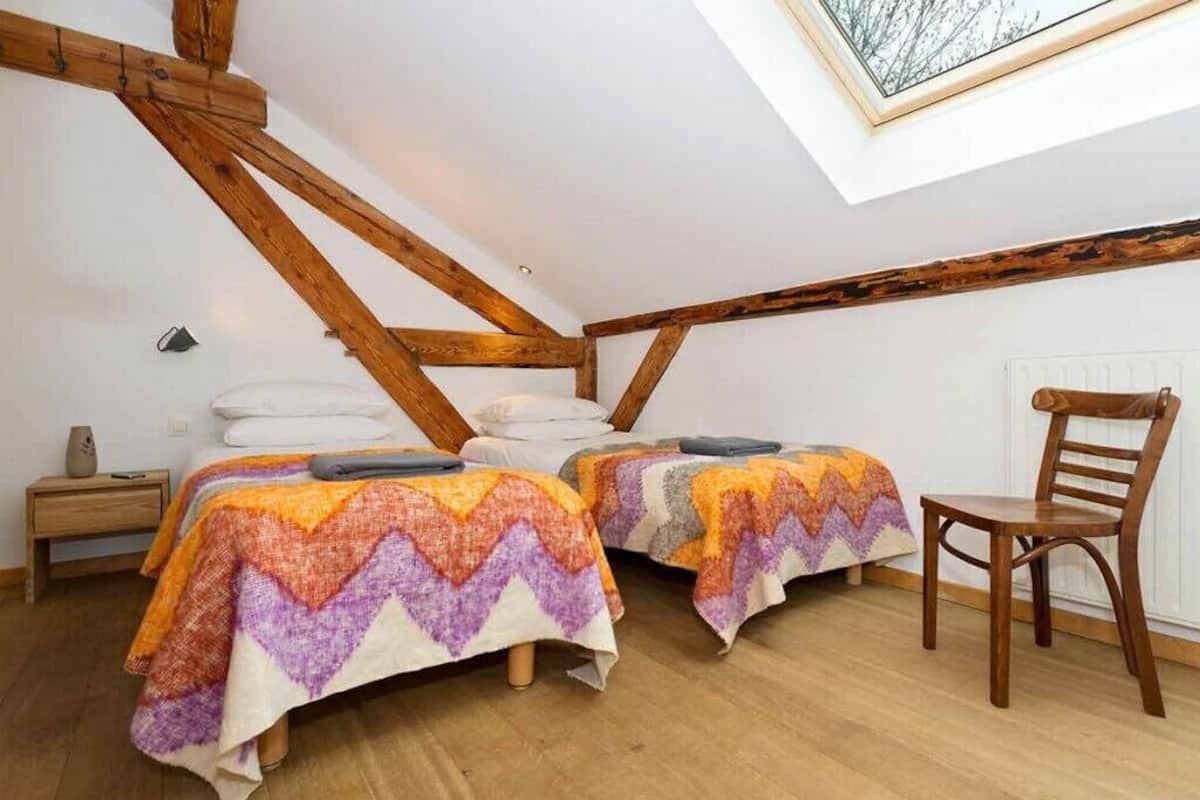 A bedroom with two beds, wooden beams, a skylight, a chair, and a nightstand.