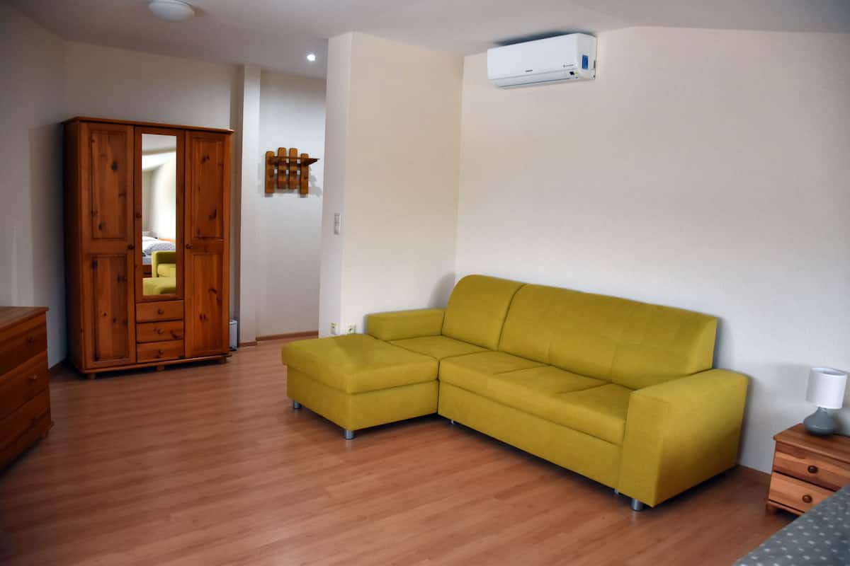 A room with a wooden wardrobe, a yellow sofa, a bedside table with a lamp, and a wall-mounted air conditioner.