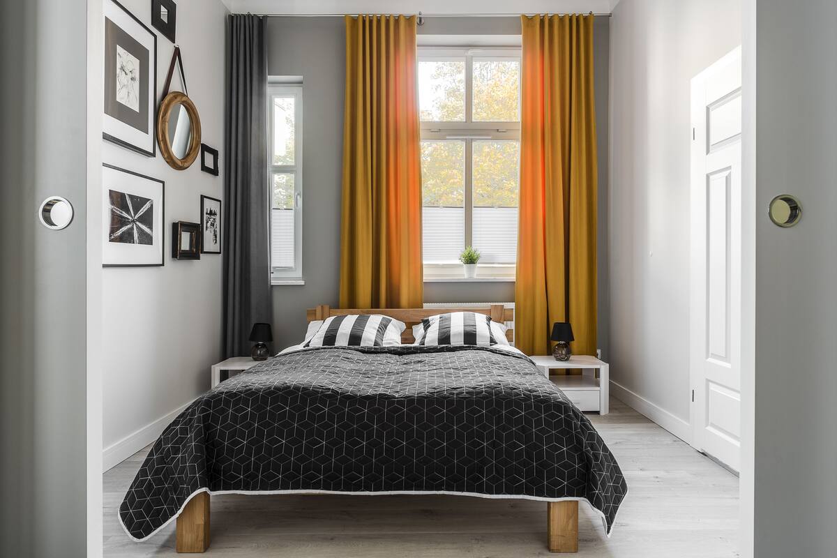 A bedroom with a bed, a bedside table, a mirror, and a window with curtains.