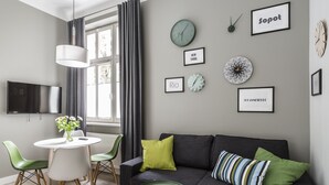 Een moderne woonkamer met een grijze bank, groene en witte stoelen, een ronde tafel en een flatscreen televisie aan de muur.