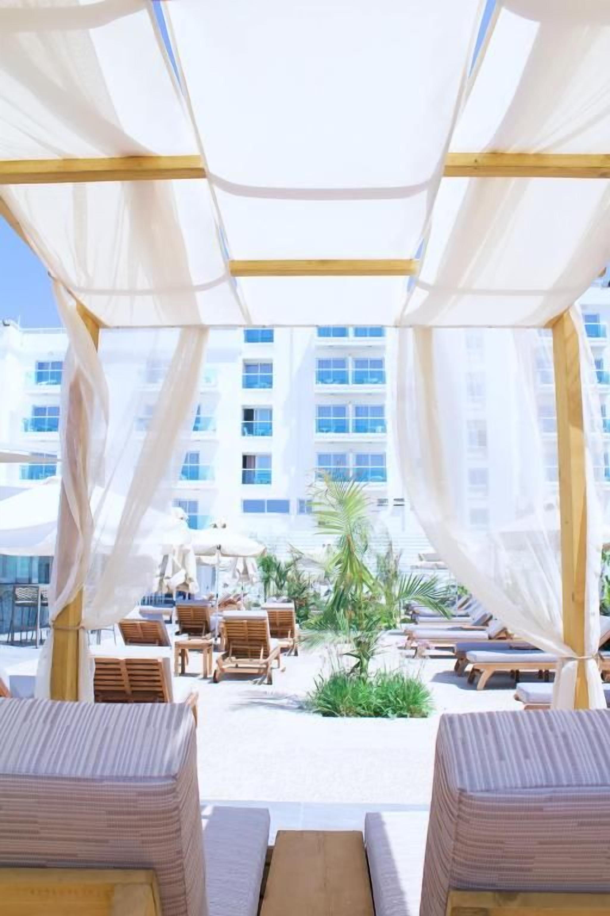 Un área de estar al aire libre con toldos blancos, muebles de madera y vista a un edificio al fondo.