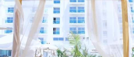 Un área de estar al aire libre con toldos blancos, muebles de madera y vista a un edificio al fondo.