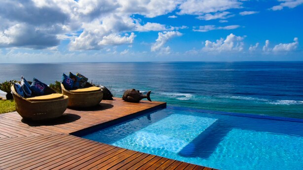 A wooden deck with a pool overlooking the ocean, two wicker chairs with cushions, and a decorative fish sculpture. - Villa Castellos (Jangamo)