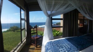A bedroom with a large window overlooking the ocean, a bed with a patterned bedspread, and a balcony with a wooden chair. - Villa Castellos (Jangamo)