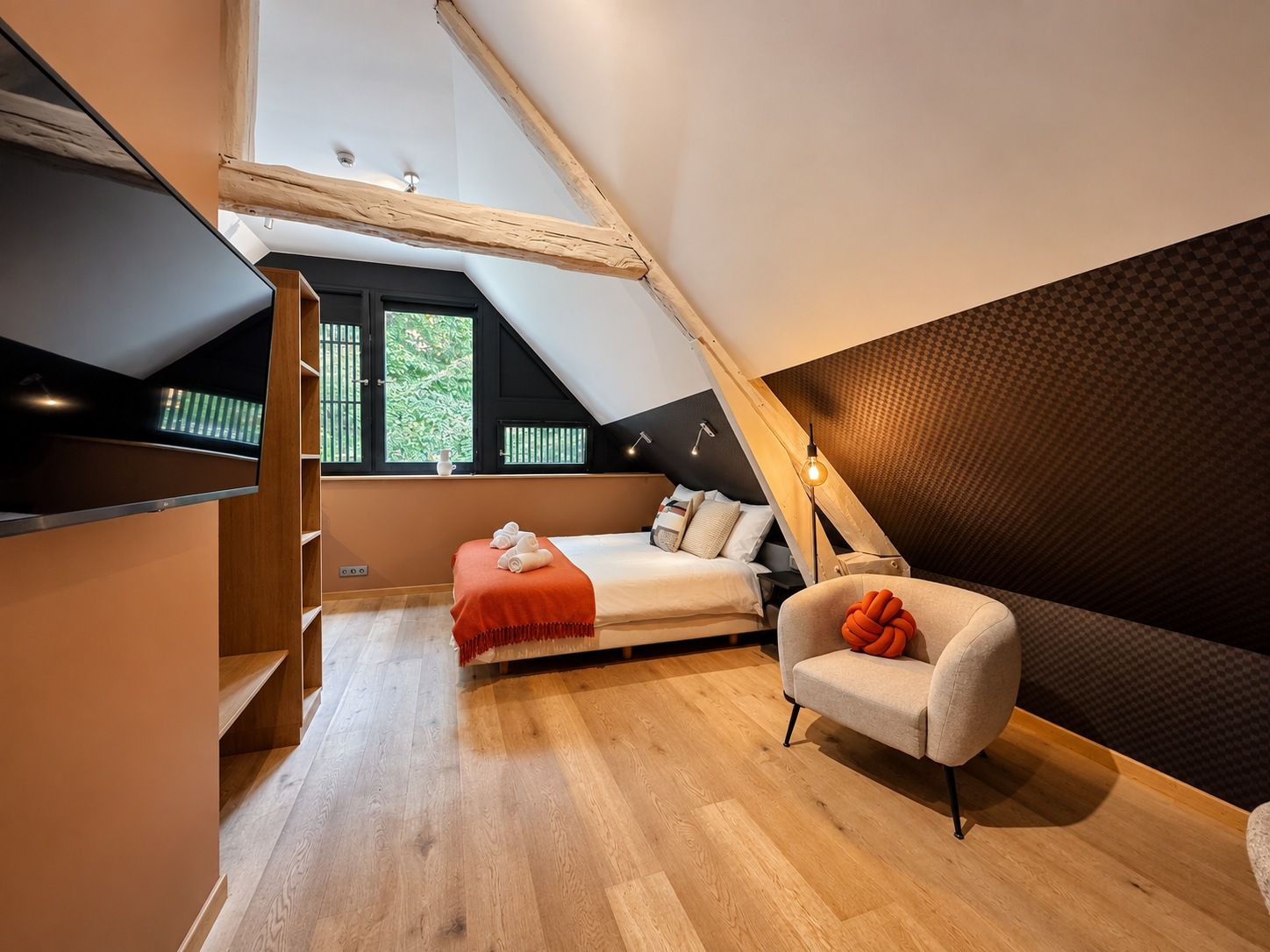 A modern loft bedroom with a sloped ceiling, a bed with a red throw, a chair with an orange cushion, and a bookshelf.