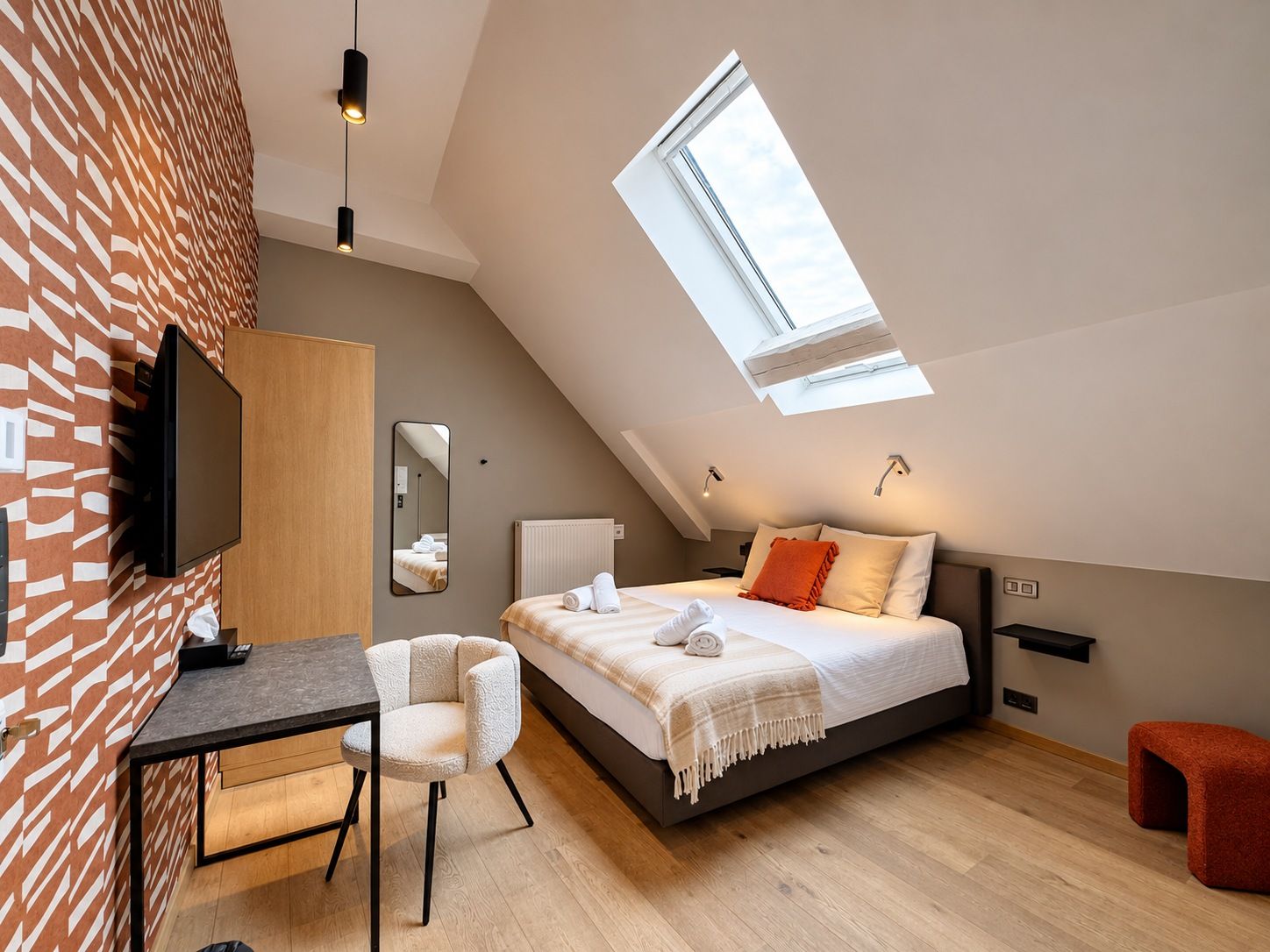 Un dormitorio moderno con techo inclinado, tragaluz, cama con almohadas, mesita de noche, una silla y un televisor.