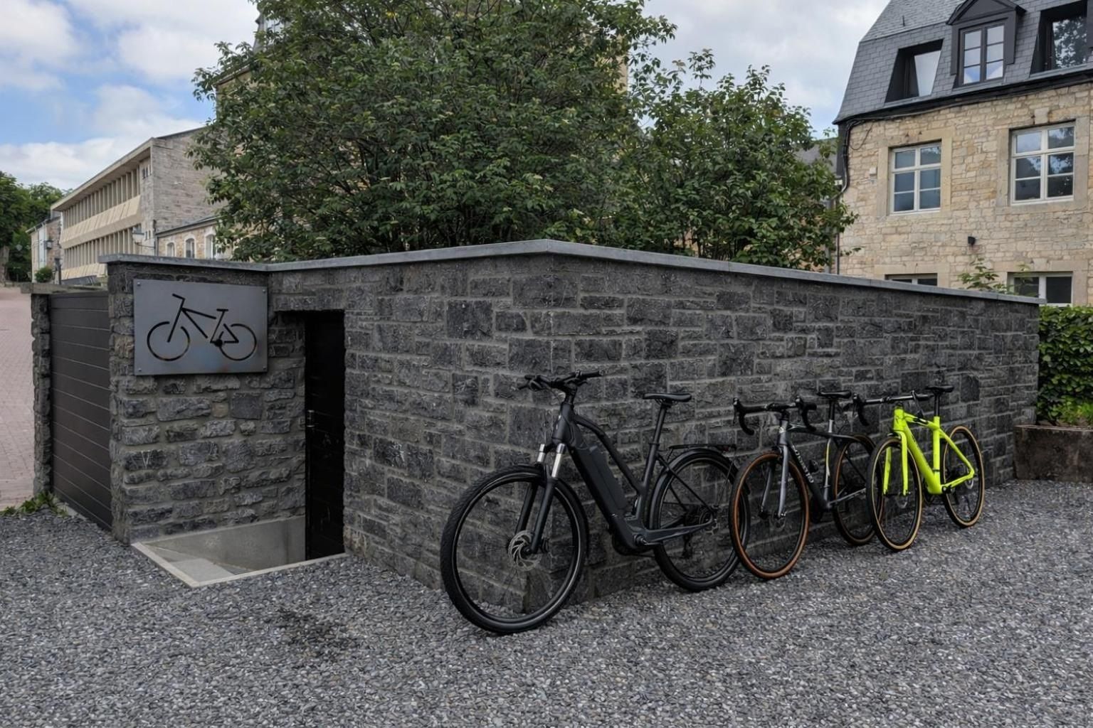 Un bâtiment doté d’une façade en pierre, d’un espace de stockage pour vélos avec un panneau indicateur et d’un sol recouvert de gravier.