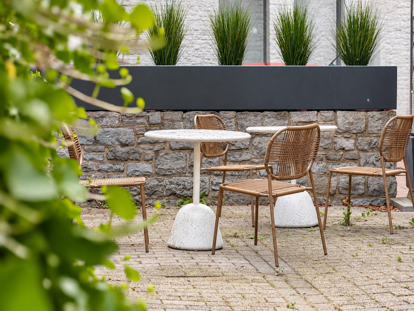 Un área de estar al aire libre con sillas de mimbre y mesas redondas, frente a una pared de piedra con plantas en macetas.