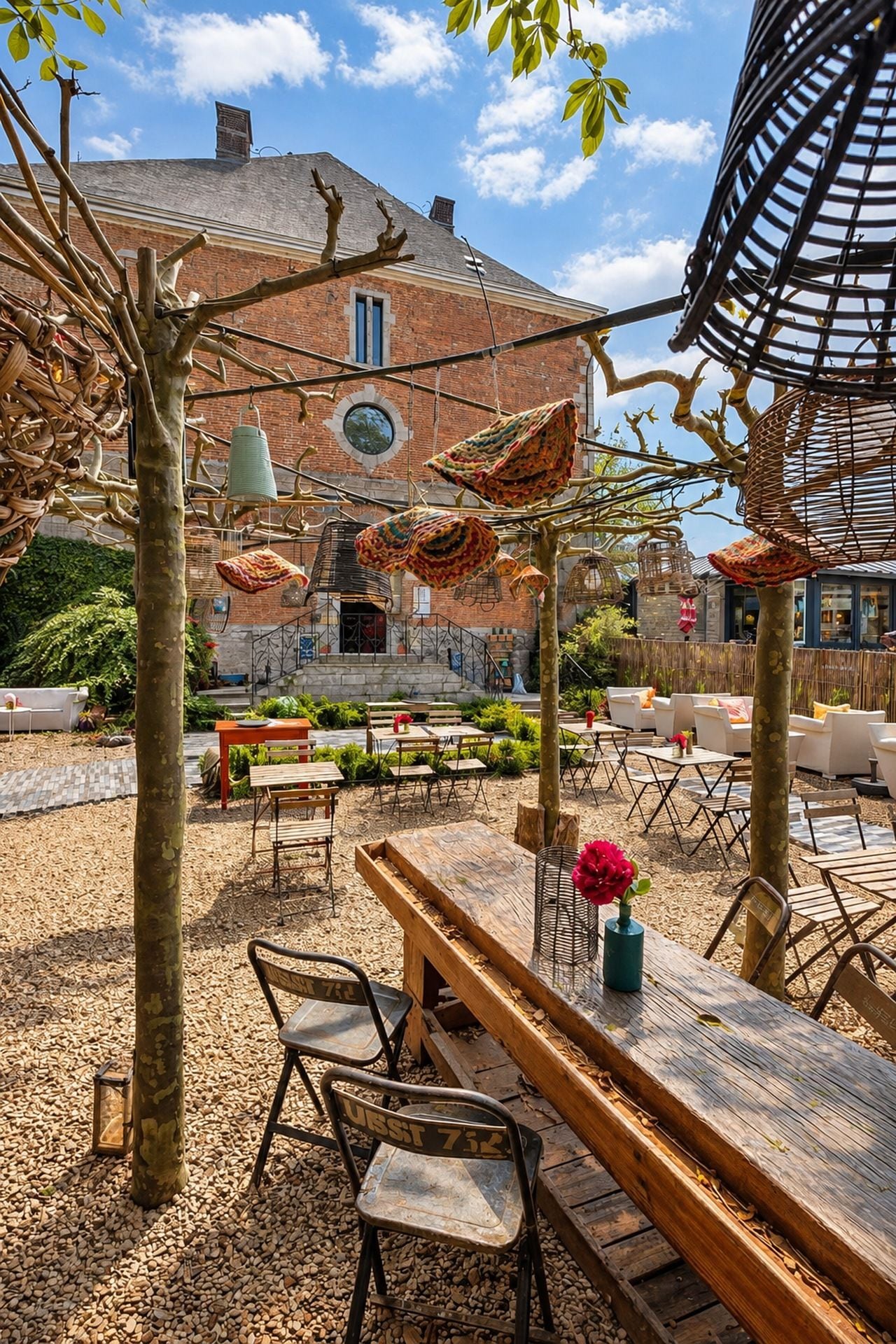 Ein Sitzbereich im Freien mit Tischen und Stühlen, ein Backsteinhaus im Hintergrund und eine Pergola mit hängenden Dekorationen.