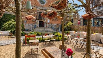 Ein Sitzbereich im Freien mit Tischen und Stühlen, ein Backsteinhaus im Hintergrund und eine Pergola mit hängenden Dekorationen.