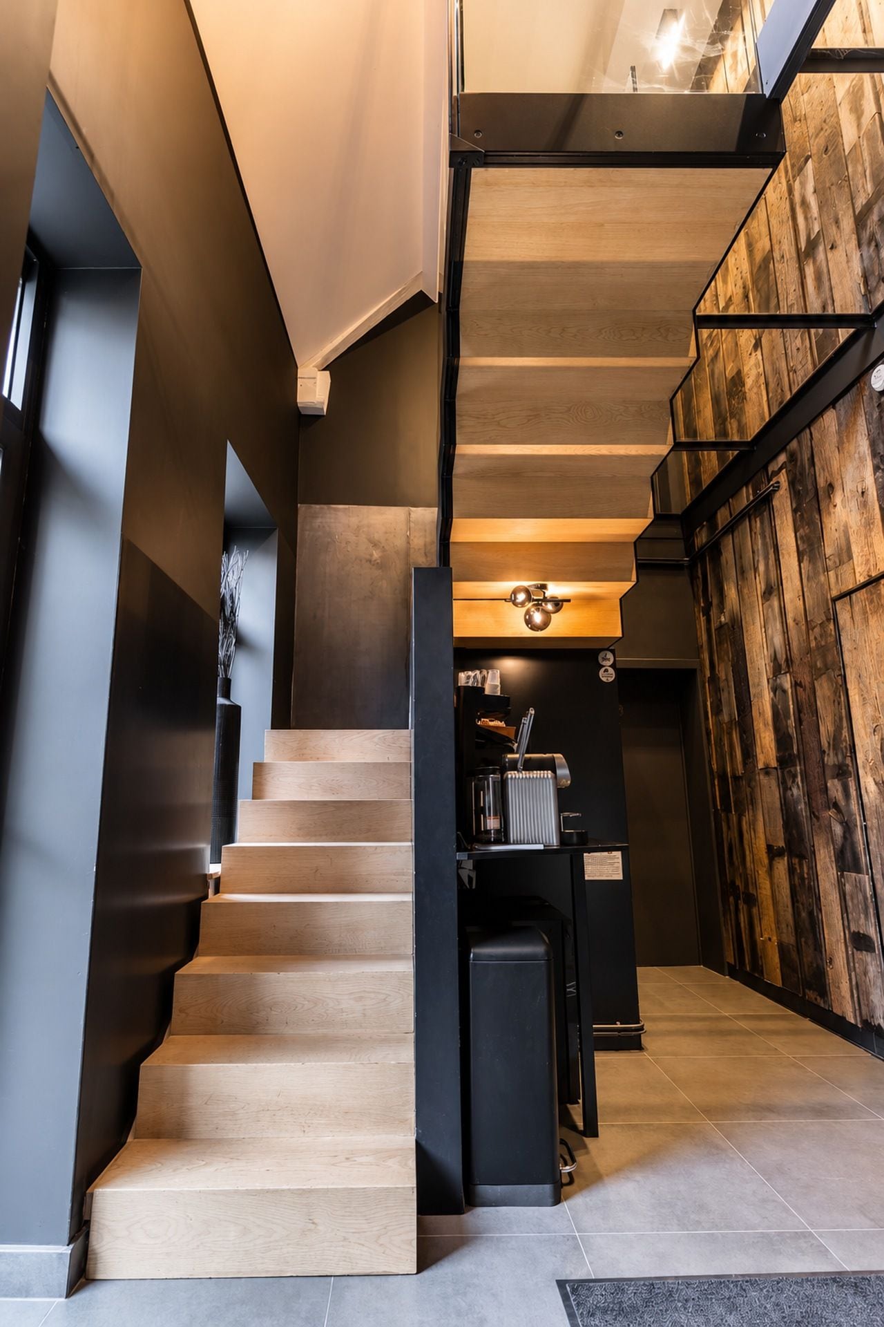 A modern staircase with wooden steps and a metal railing, leading to a room with a wooden wall and a coffee machine.