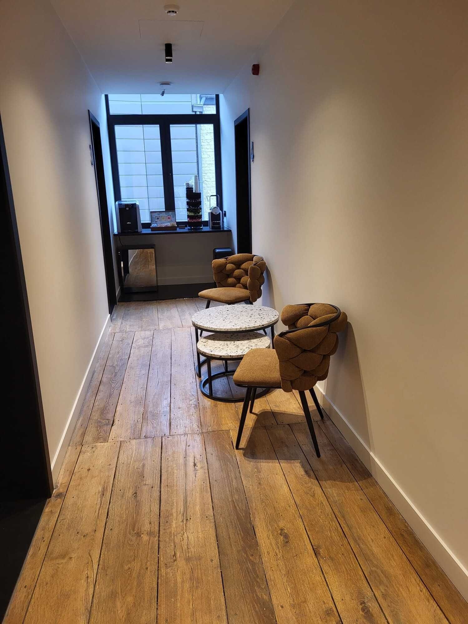 A narrow hallway with wooden flooring, a round table with chairs, and a window with a view of the outside.