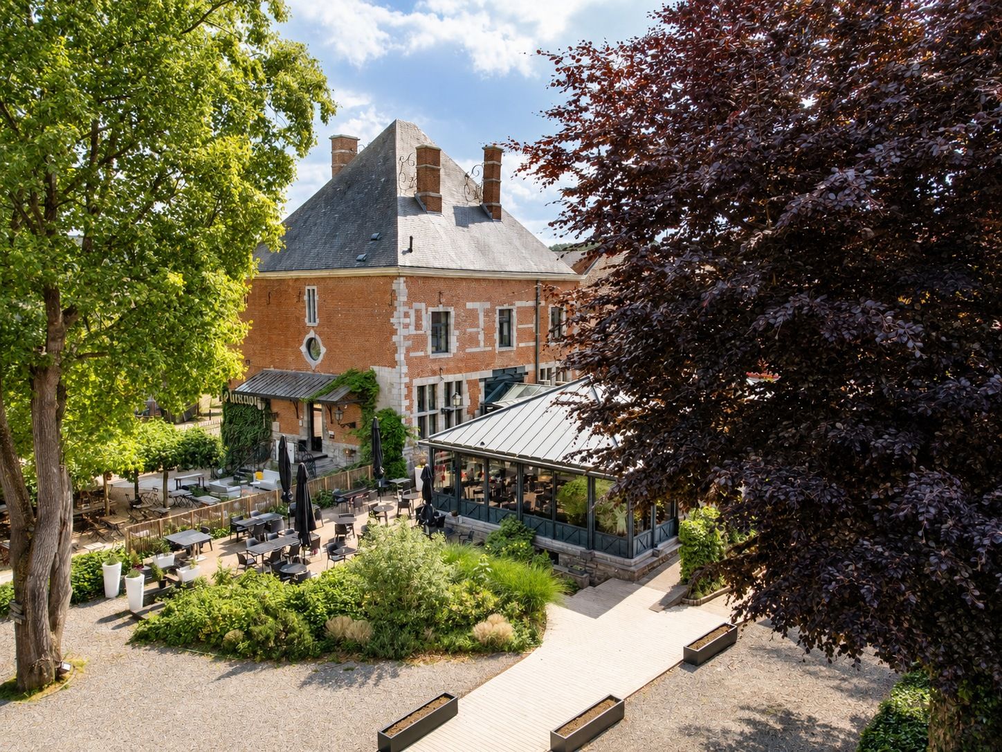 A courtyard with outdoor seating, a brick building with a glass conservatory, and a tree-lined path.