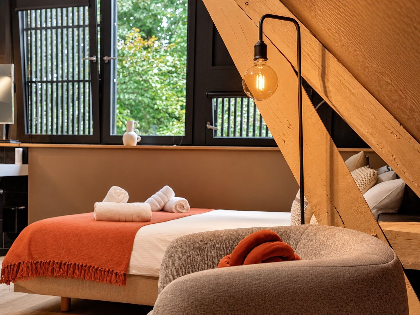 A bedroom with a bed, a wooden sloped ceiling, a window with a view of greenery, and a hanging light fixture.