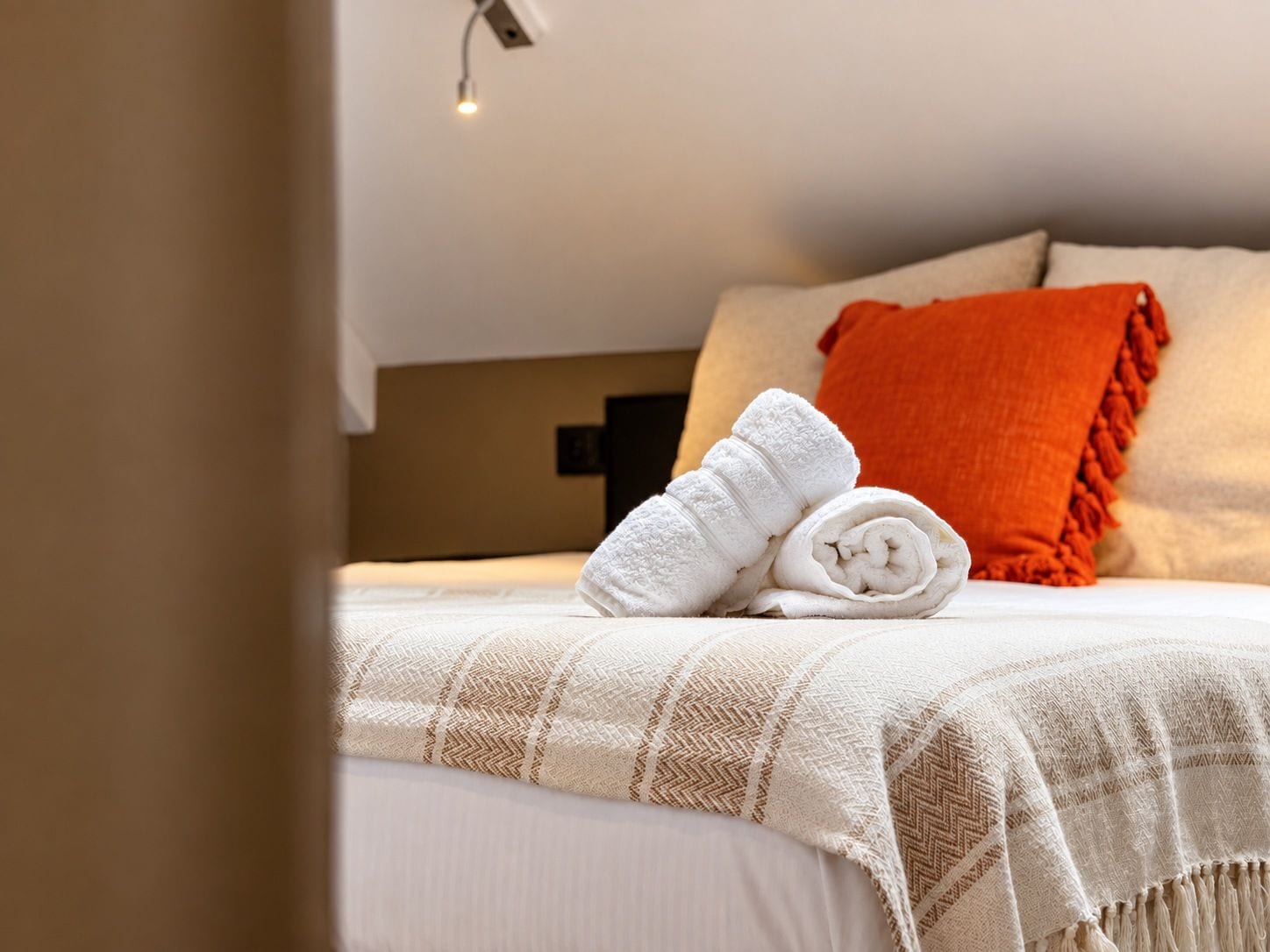Una cama bien hecha con una colcha blanca y marrón a cuadros, una toalla blanca enrollada y una almohada naranja.