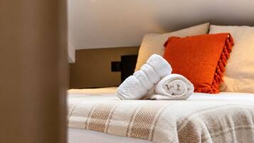 Una cama bien hecha con una colcha blanca y marrón a cuadros, una toalla blanca enrollada y una almohada naranja.