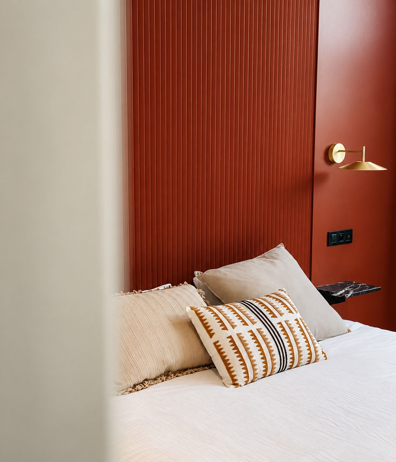 Una cama con ropa blanca y almohadas estampadas, pared texturizada de color rojo y lámpara dorada.