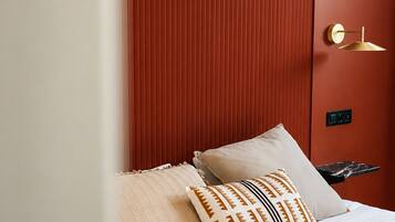 Una cama con ropa blanca y almohadas estampadas, pared texturizada de color rojo y lámpara dorada.