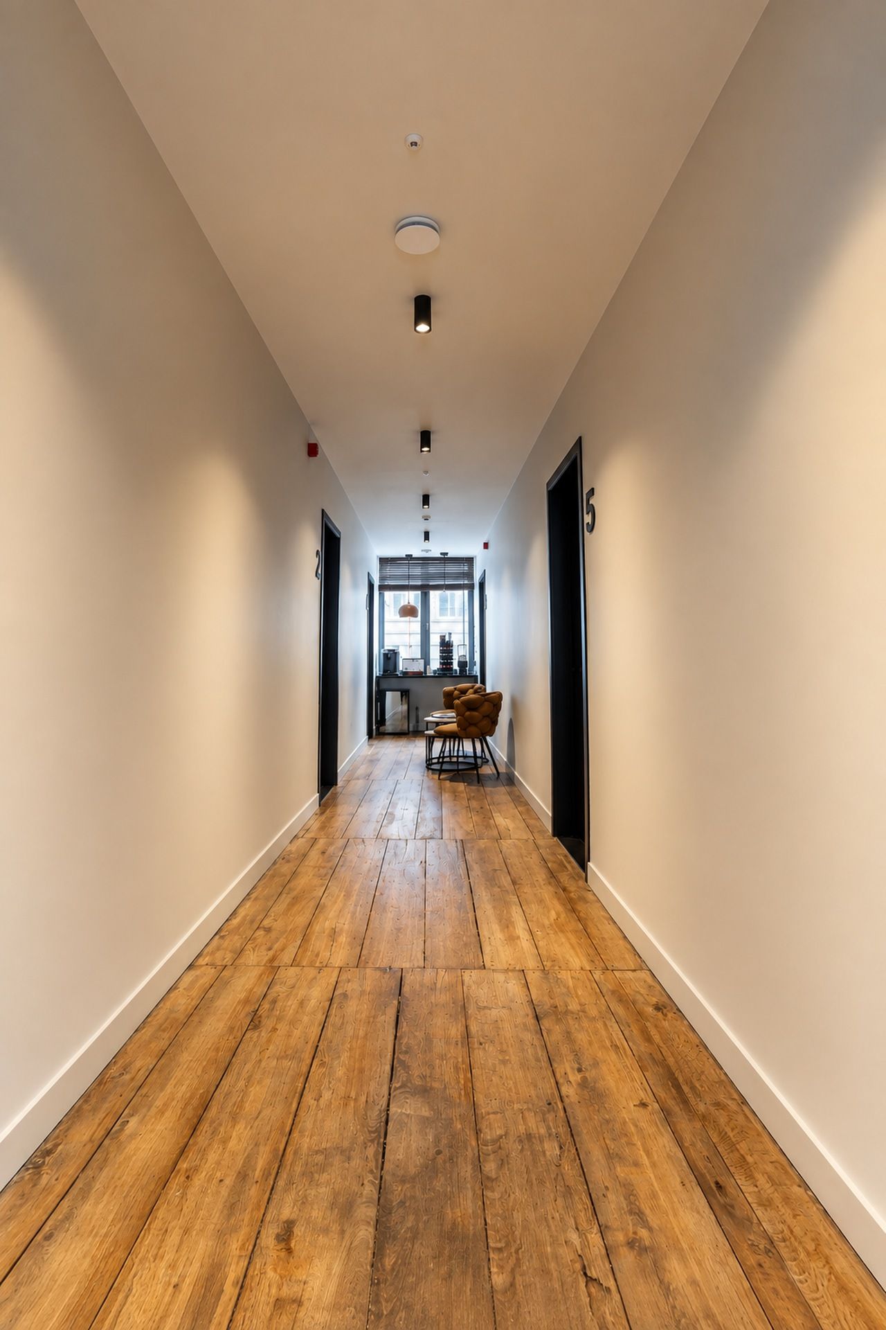 Un couloir doté d’un plancher en bois, de murs blancs et de portes de part et d’autre.