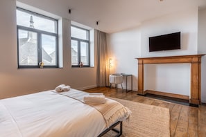 A hotel room with a large bed, a fireplace, a TV, and a view of a building through the windows. - Hôtel Le Manoir (Marche-en-Famenne)