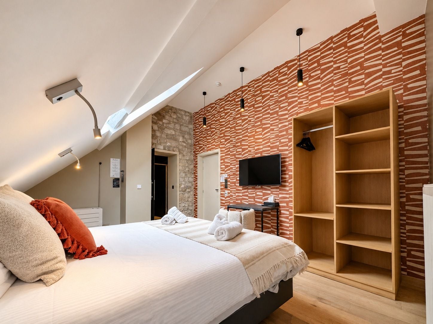 Quarto moderno com teto inclinado, cama com roupa de cama branca, guarda-roupa de madeira e televisão na parede.