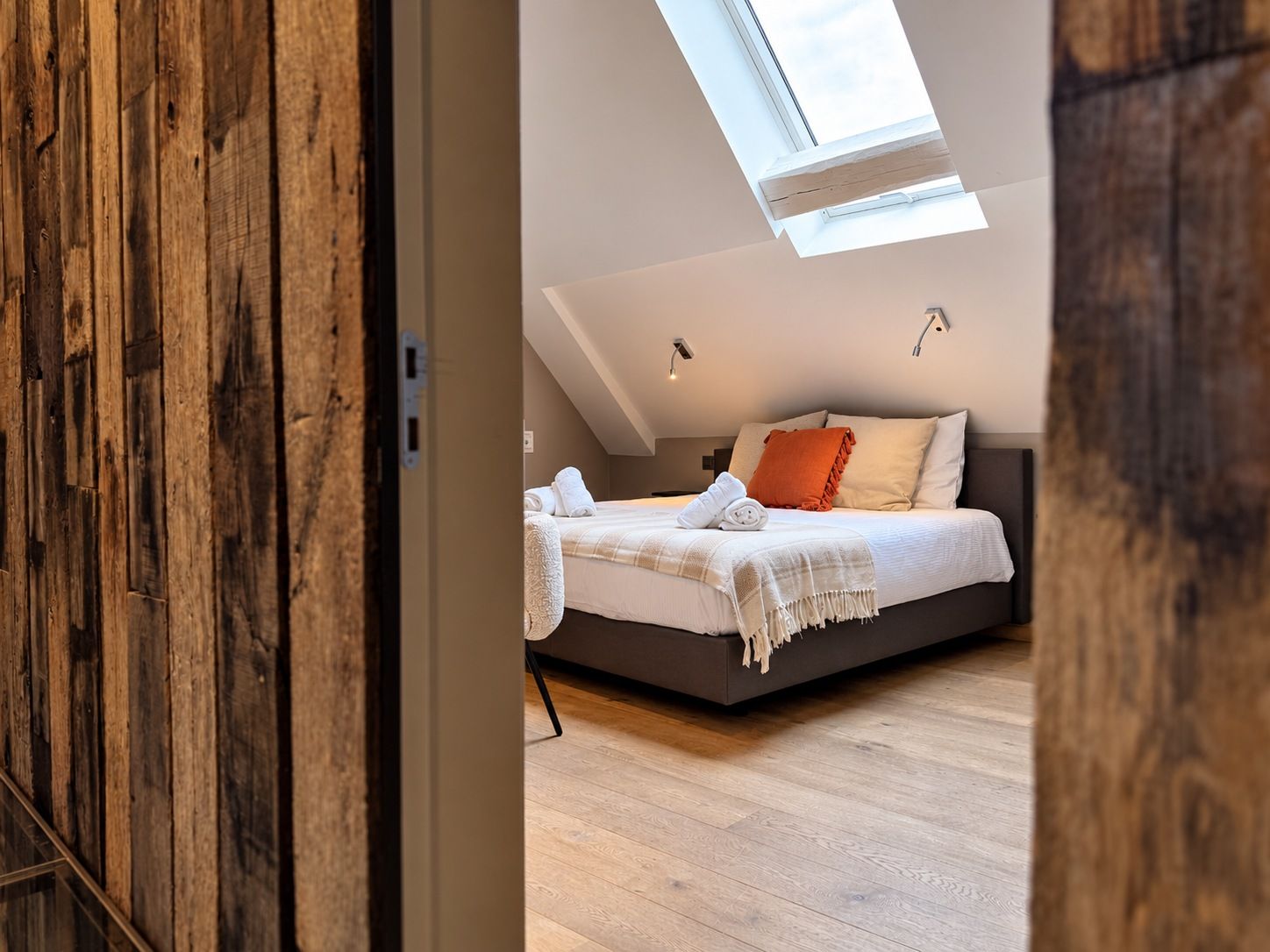 A bedroom with a sloped ceiling, a skylight, a bed with pillows and a blanket, and a wooden floor.