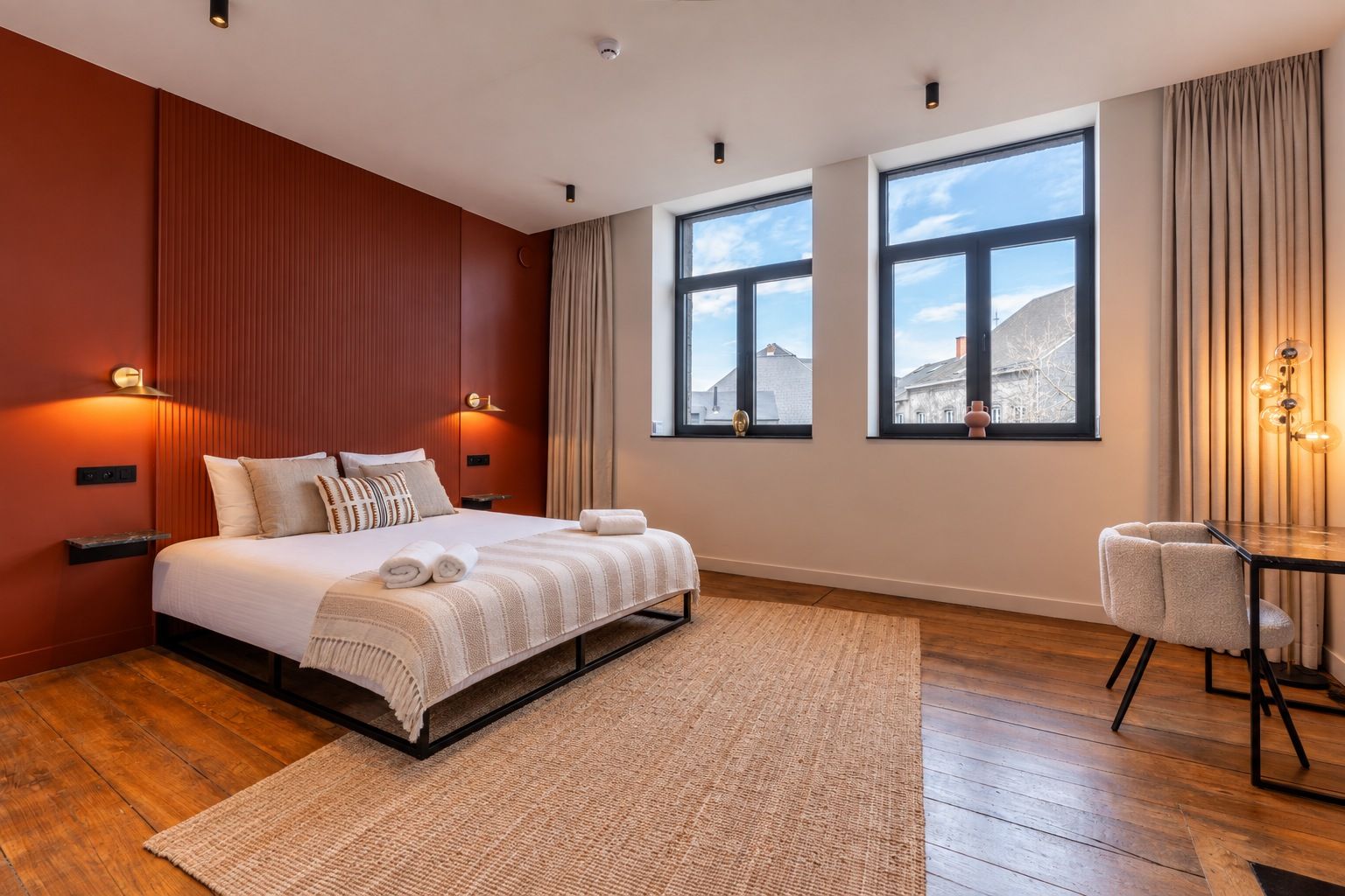 Une chambre d’hôtel moderne avec un grand lit, une petite table et deux fenêtres donnant sur la ville.