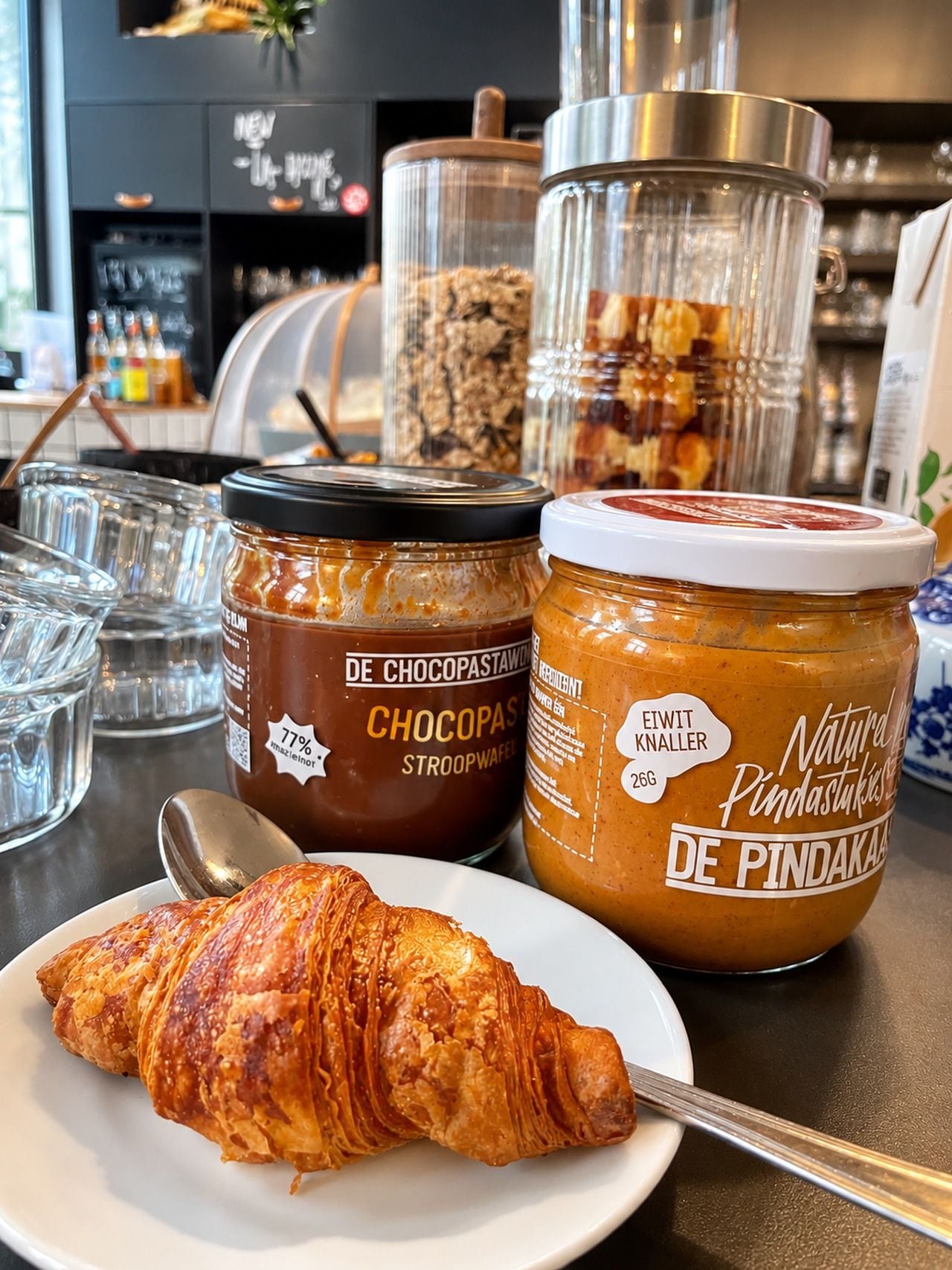 Un croissant sur une assiette, deux pots de pâte à tartiner et une cuillère.