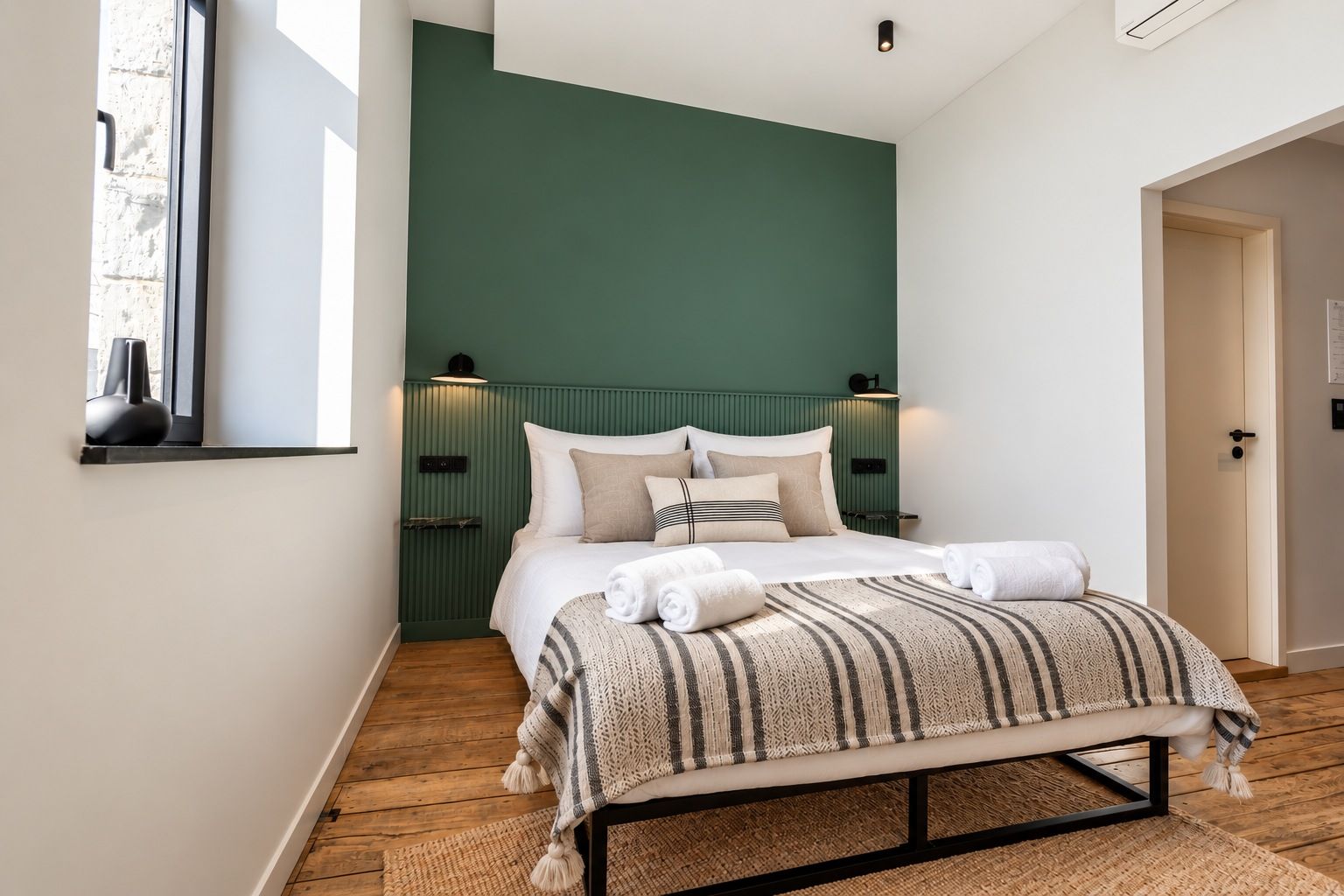 Un dormitorio con cama, ventana, puerta y una lámpara fijada a la pared.