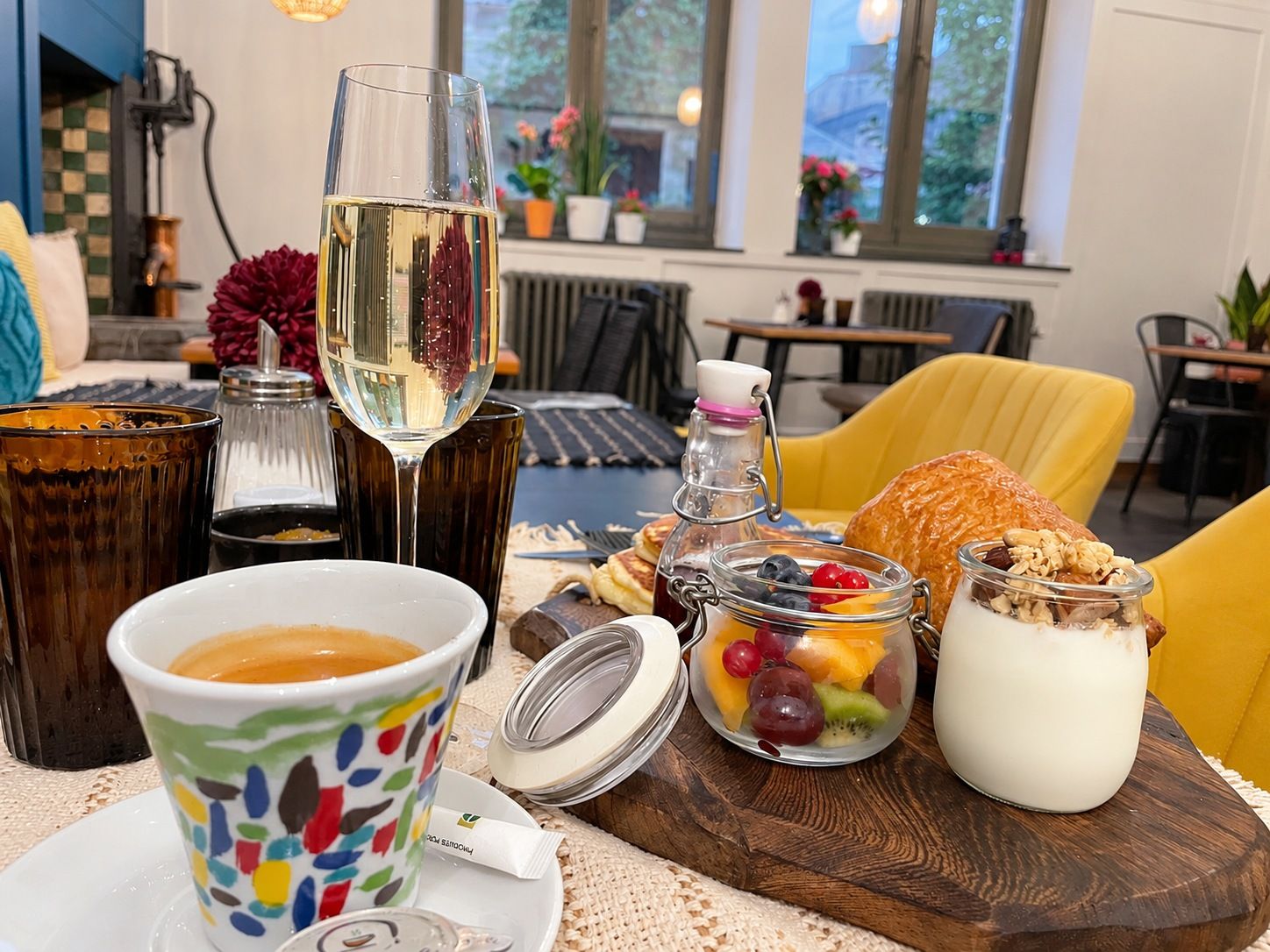 A breakfast setting with a cup of coffee, a glass of white wine, a jar of fruit, a pastry, and a glass of milk on a wooden board.