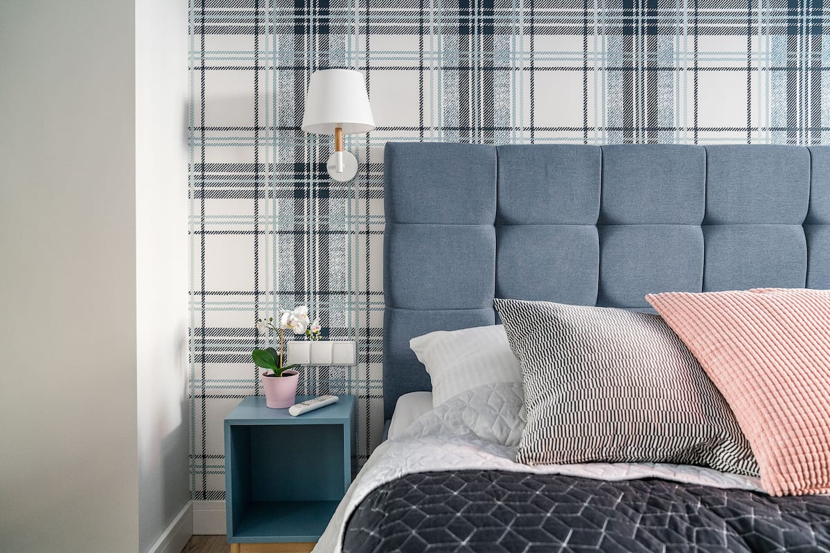 A bedroom with a plaid-patterned wall, a blue headboard, a bedside table with a lamp, and a potted plant.