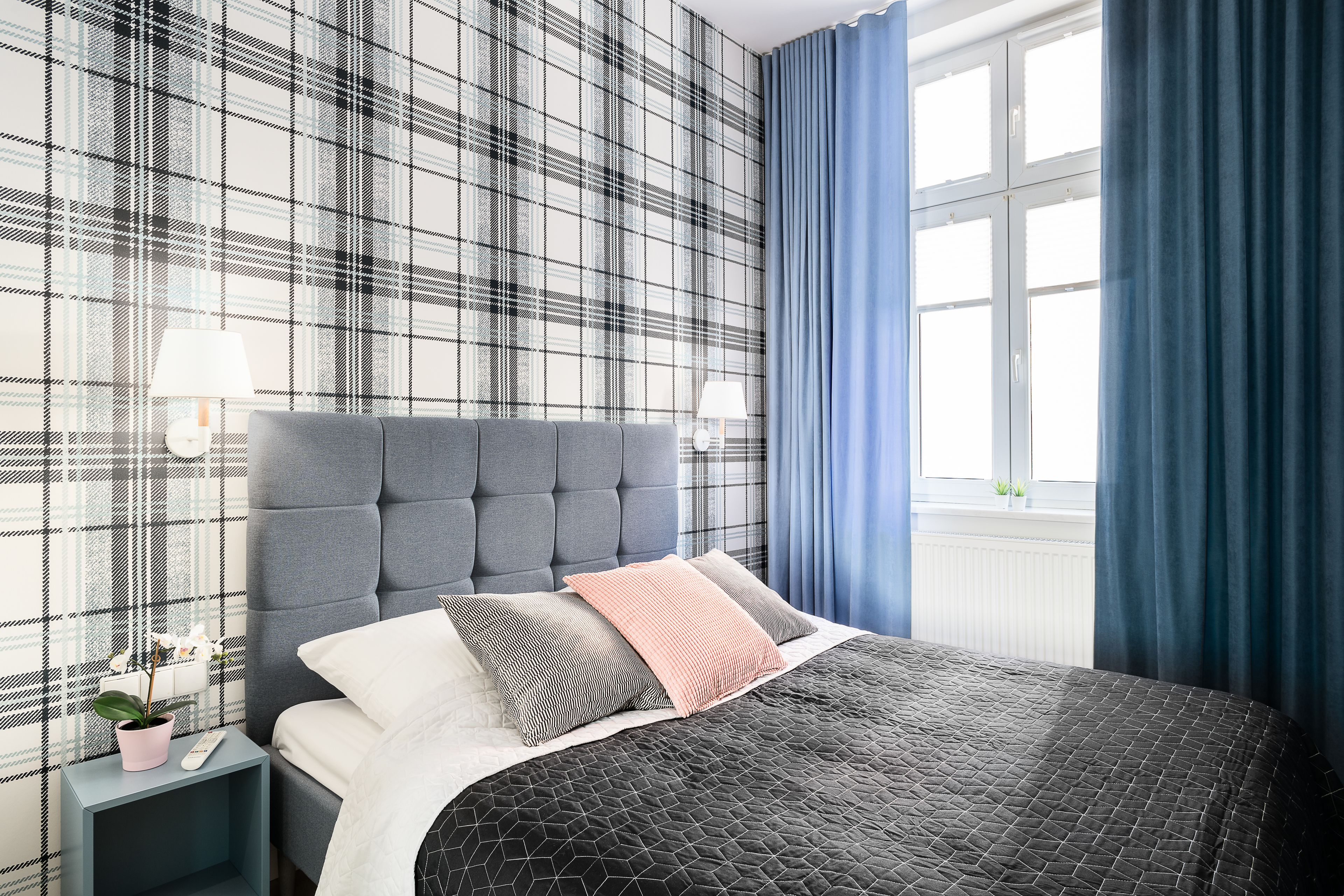 A bedroom with a plaid wallpaper, a tufted headboard, a bed with a quilted cover, a bedside table with a lamp, and a window with blue curtains.