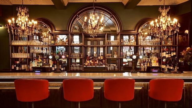 A bar area with three red bar stools, a mirrored wall, and a variety of bottles and glasses on shelves. - The Bedford Balham (London)