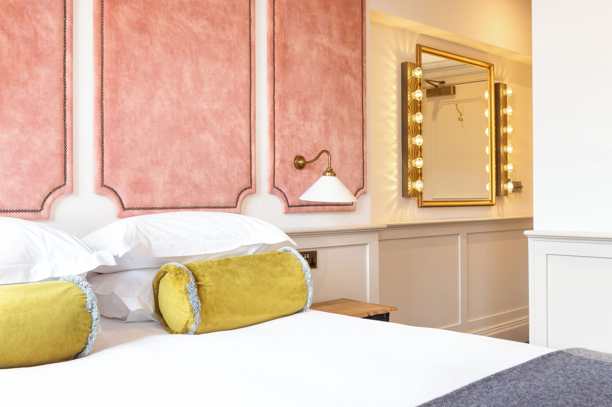 A hotel room with a bed, a nightstand, a mirror with lights, and a wall-mounted lamp.