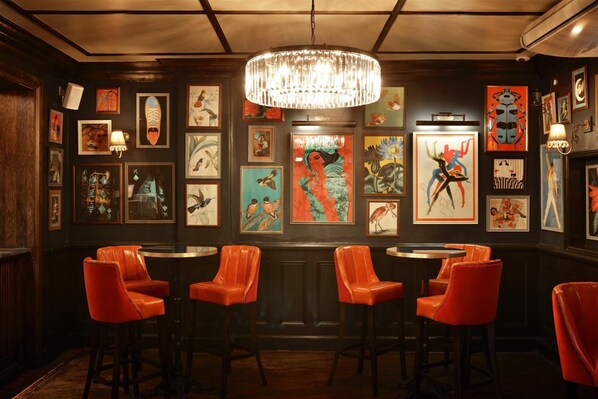 A bar area with red leather bar stools, a chandelier, and various framed artworks on the walls. - The Bedford Balham (London)