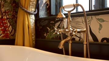 Una bañera antigua con grifo y manguera de latón, frente a una pared con papel tapiz floral.
